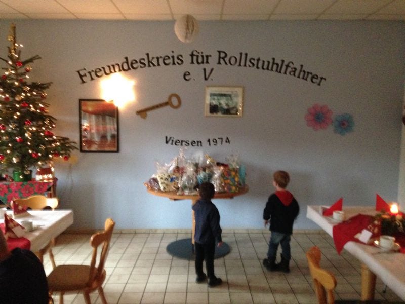 Bild: Impression der Weihnachtsfeier von 2017. Auf dem Bild wird eine geschmückte Wand mit einem Weihnachtsbaum davor gezeigt. Es stehen zwei Kinder vor einem Geschenke-Tisch.
